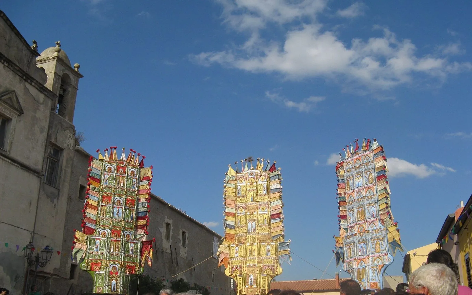 I tre candelieri di Nulvi, alti tabernacoli decorati nei colori verde, oro e azzurro con bandierine, sollevati contro il cielo blu accanto a un'antica chiesa
